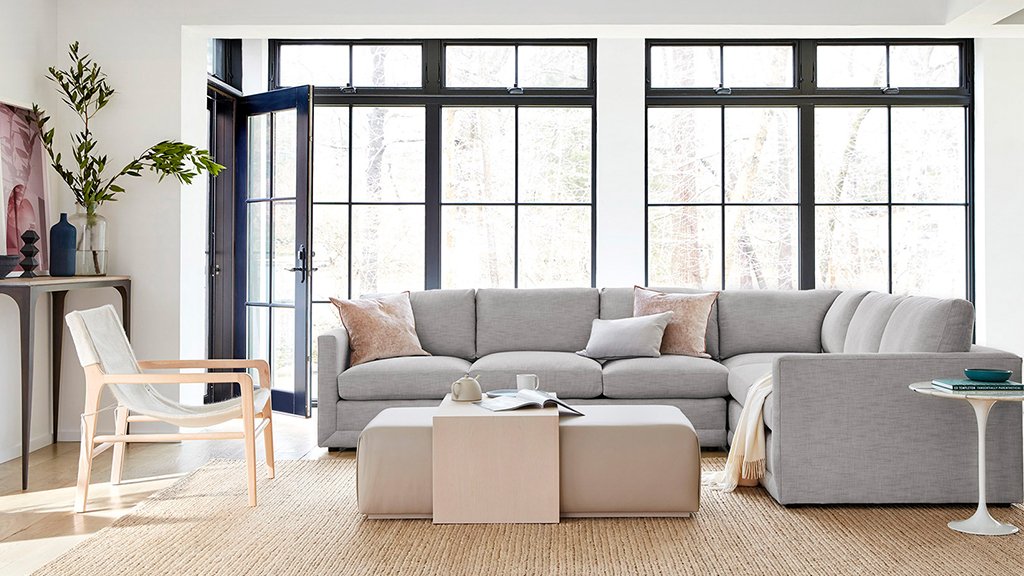 A modern living room with large black-framed windows, light gray sectional sofa, beige rug, and a minimalist chair with wooden frame. The room includes a matching pair of light gray ottomans serving as a coffee table, and a small side table with a vase and a book.