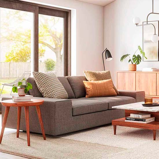 A modern living room featuring a gray sofa adorned with patterned cushions, a round wooden side table with a potted plant, and a rectangular coffee table. A floor lamp stands nearby, while large windows with a backyard view allow natural light to flood the space.