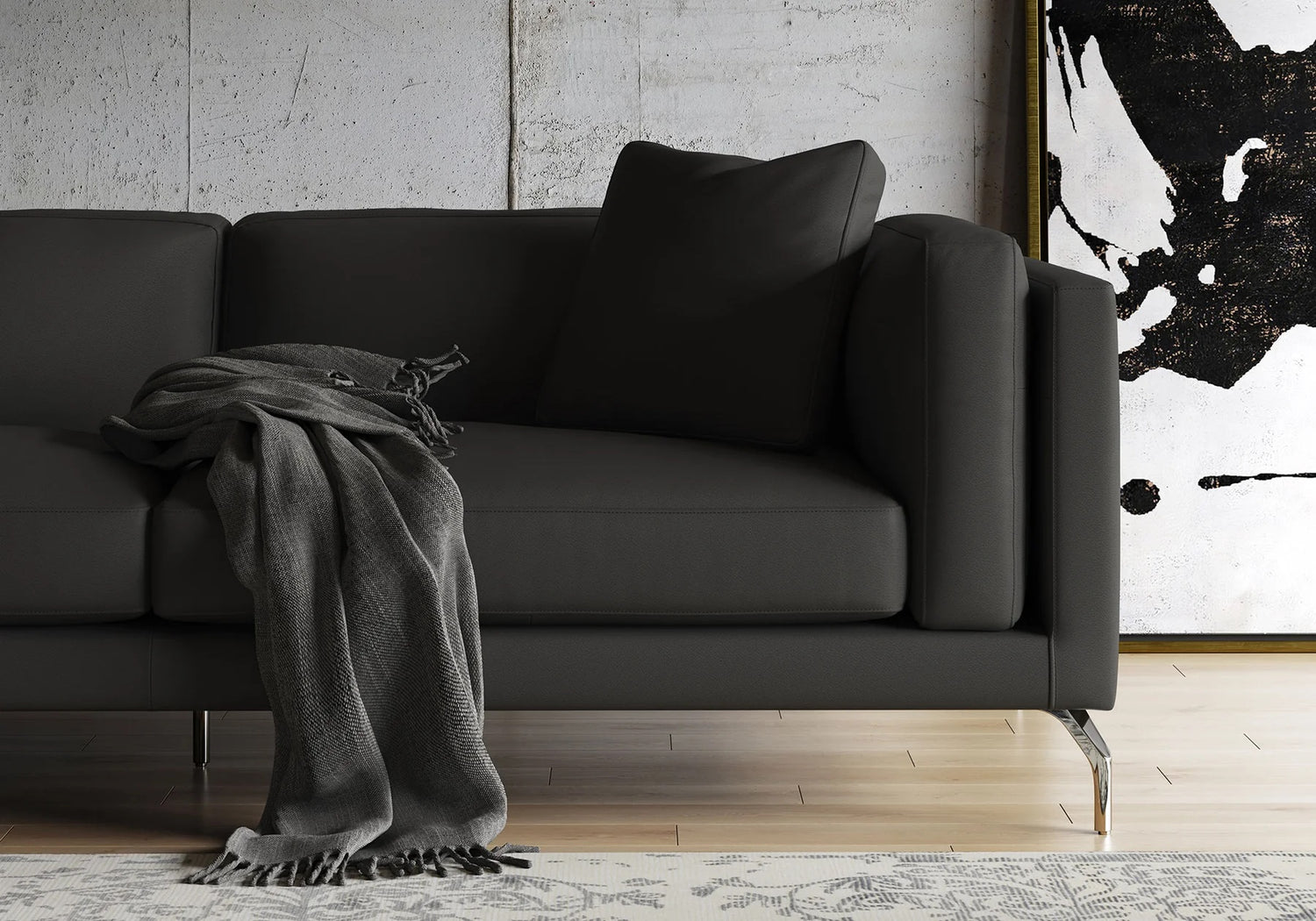 A modern black sofa with clean lines is placed in a living room with a large abstract black and white painting behind it. A dark grey throw blanket is draped over the arm of the sofa. The room features a light-colored wooden floor and a patterned rug.