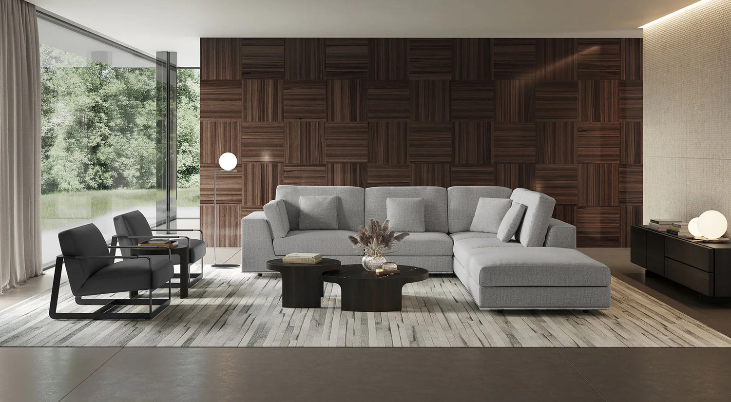 A modern living room features a large, gray sectional sofa with cushions, a dark wooden coffee table, two black armchairs, and a matching sideboard. The back wall is covered in a textured wooden panel design, and a large window offers a view of lush greenery.