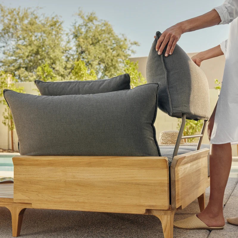 A person in a white outfit arranges the gray cushions on a wooden outdoor lounge chair. The chair is set on a patio near a pool with trees and a wall in the background. The scene appears bright and sunny.