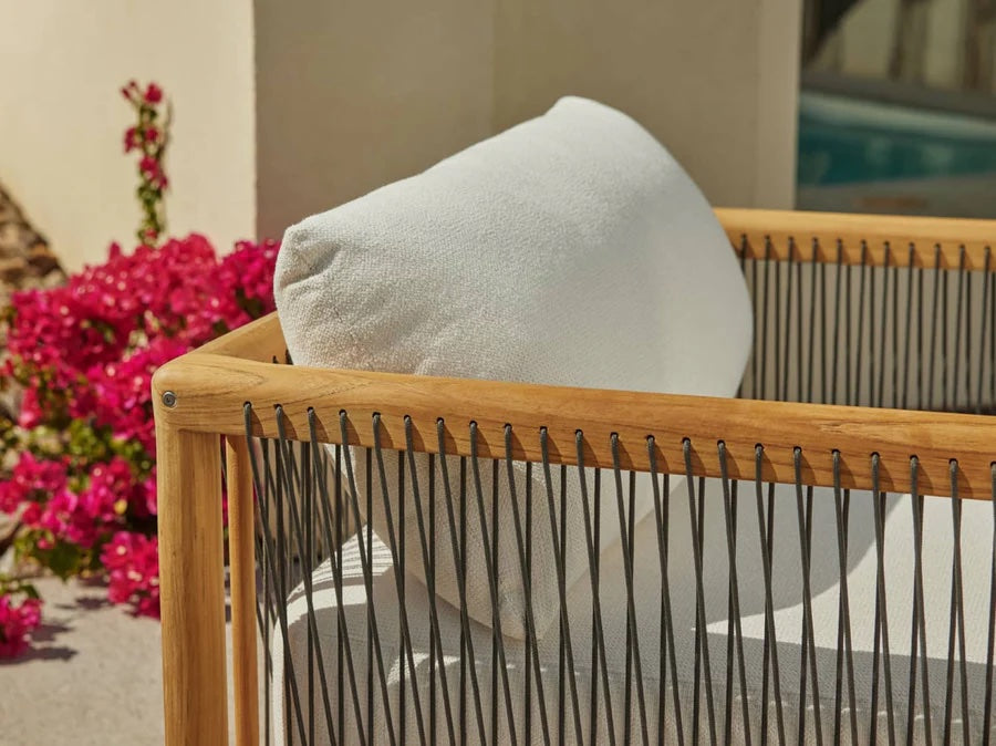 A close-up of a modern outdoor chair with a wooden frame and black rope detailing, featuring a white cushion and pillow. The chair is placed beside a vibrant pink flowering plant and near a light-colored wall, with a glimpse of a pool in the background.