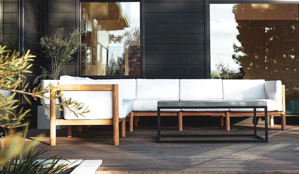 Modern outdoor patio with light wooden furniture and white cushions. The area features a large corner sofa, an armchair, and a rectangular black coffee table set on a wooden deck, with large windows in the background and some greenery.