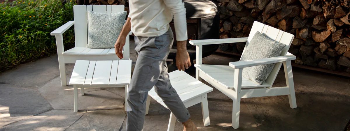 A person in a white long-sleeve shirt and gray pants walks barefoot on a patio. The patio has white wooden outdoor furniture, including a chair, loveseat, and table, with gray cushions. There is a stack of firewood in the background.