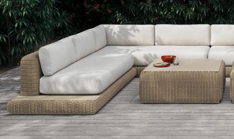 An outdoor seating area featuring an L-shaped wicker sofa with white cushions and a matching wicker coffee table. The setup is on a wooden deck surrounded by lush greenery. A couple of red bowls are placed on the coffee table.