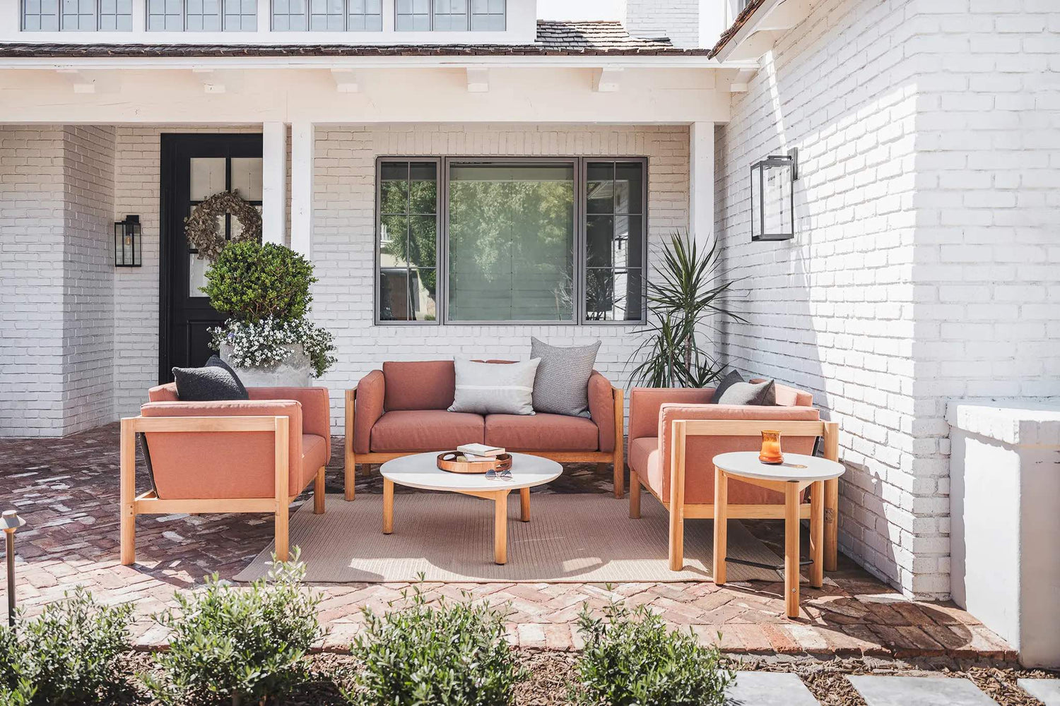 A cozy patio features a modern outdoor seating arrangement with a peach-colored sofa, two matching armchairs, and round white tables. The space is accented with green plants and white brick walls, creating a serene atmosphere for relaxation and social gatherings.