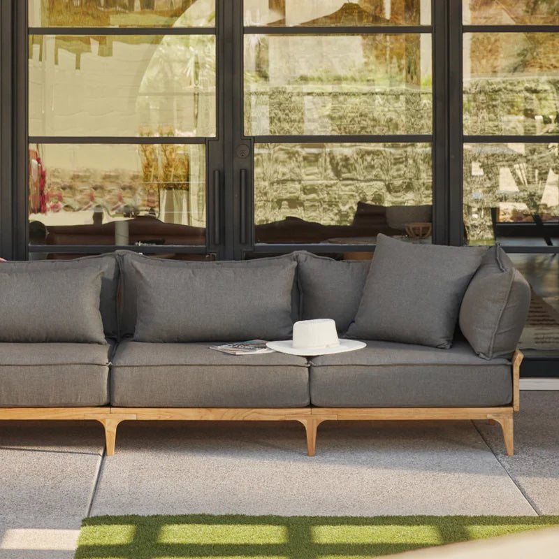 A modern outdoor patio setup featuring a cushioned wooden sofa with dark gray cushions. A white hat and a magazine are placed on the sofa's armrest. The background shows large glass doors reflecting a rustic stone wall.