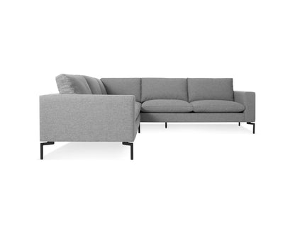 The New Standard Sofa Sectional Sofa features a gray L-shaped design with clean, modern lines and slender black metal legs. Its plush cushions enhance the comfort of its sleek profile. Positioned against a white background, it highlights its contemporary style.