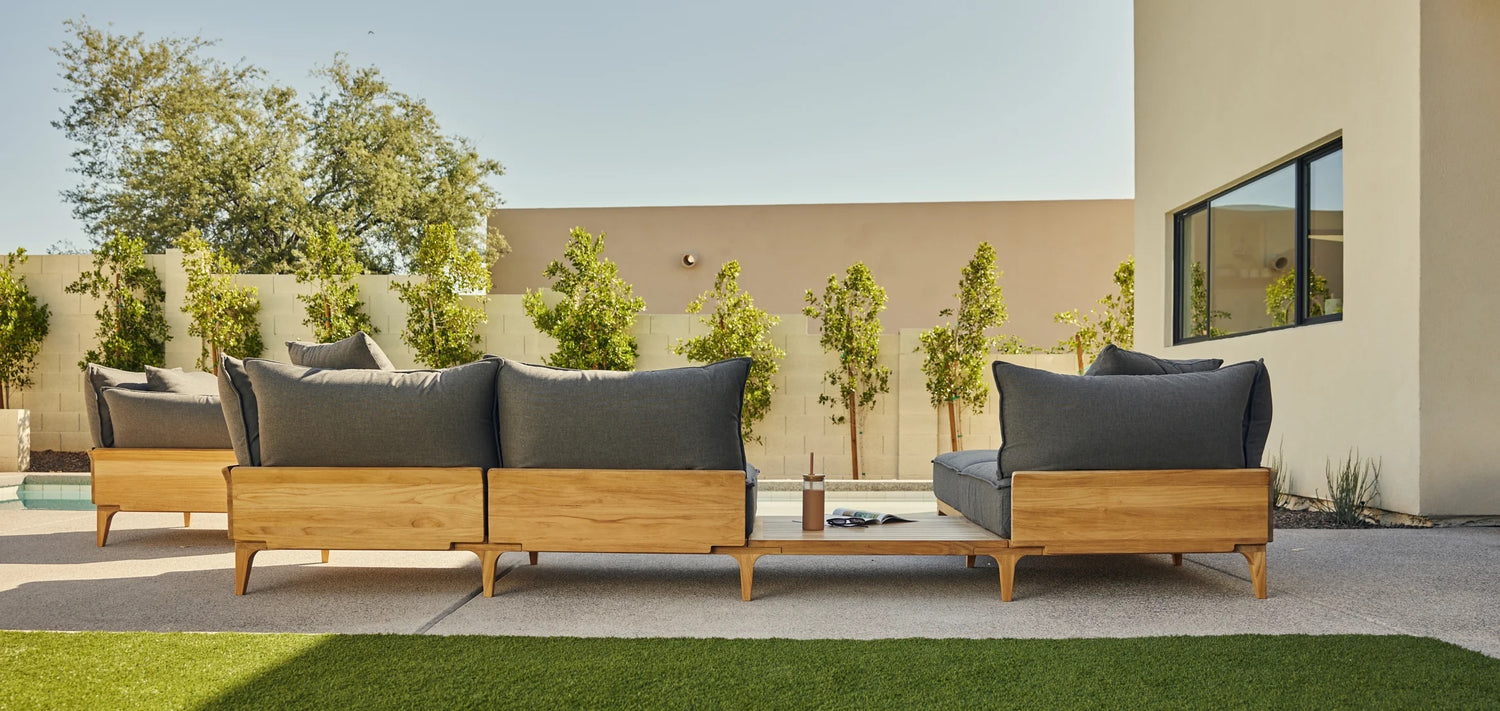 A modern outdoor seating area with wooden-framed sofas and gray cushions facing a pool. Behind the seating, there is a beige wall with trimmed bushes and trees. The area is well-lit with natural sunlight, and the grass lawn is partially visible in the foreground.