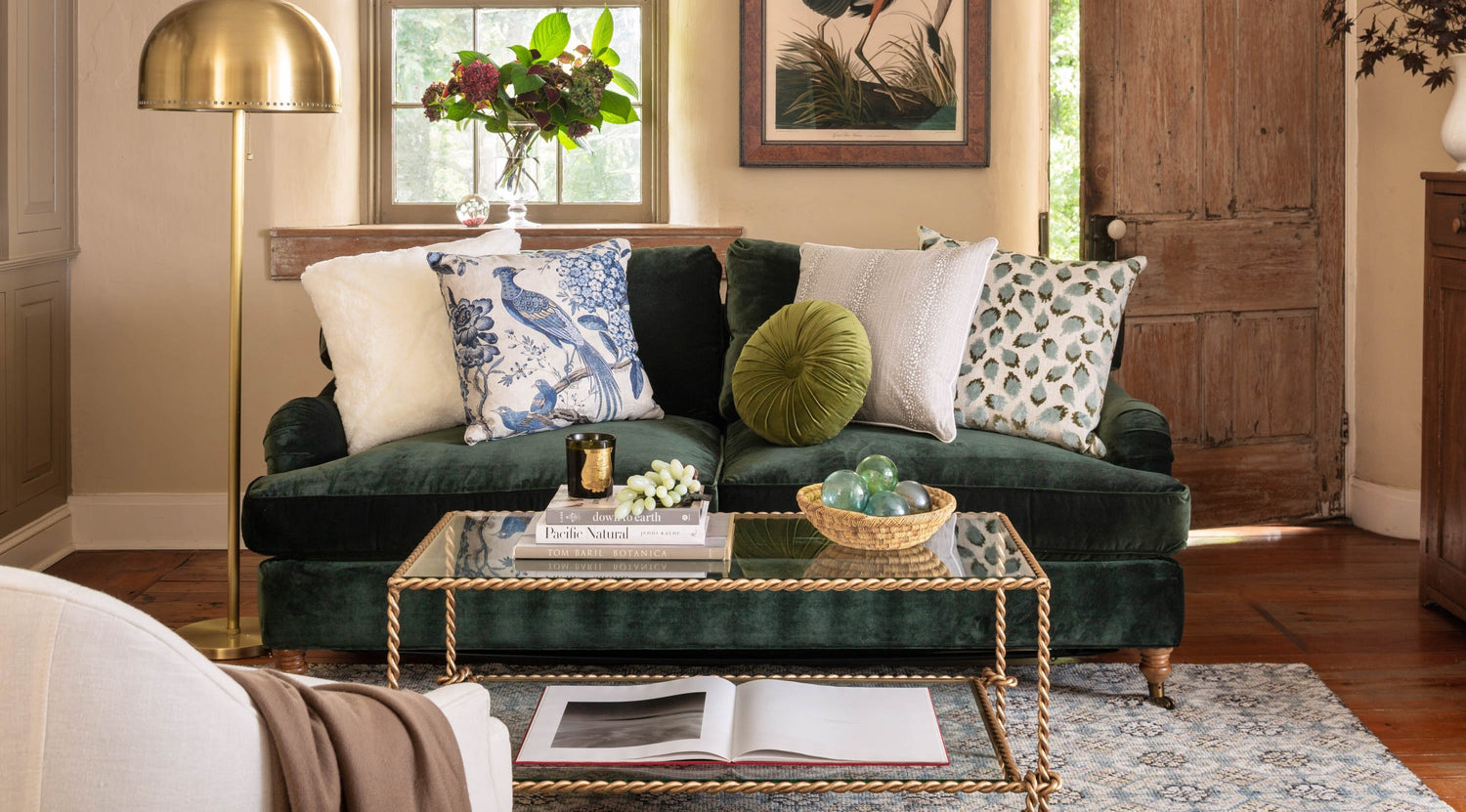 A cozy living room features a cushioned armchair with a green floral pattern, a dark green sofa with decorative pillows, and a wooden coffee table with a vase of pink flowers. Blue and white decorative plates adorn the wall, and a side table holds pastel flowers.