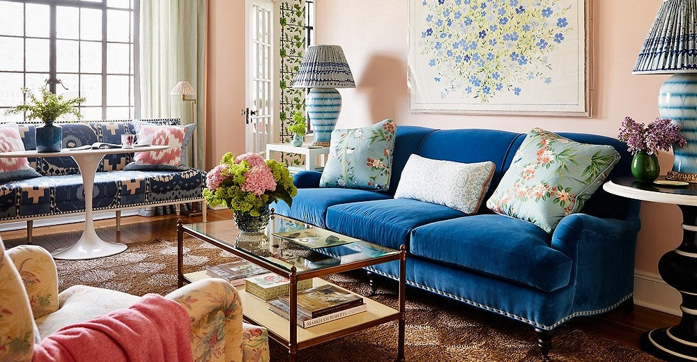 A bright living room features a blue sofa with a patterned pillow, a white and blue accent chair, and a table with blue and white striped chairs. There are plants, decorative vases, books, and a modern line-art piece on the wall with large windows in the background.