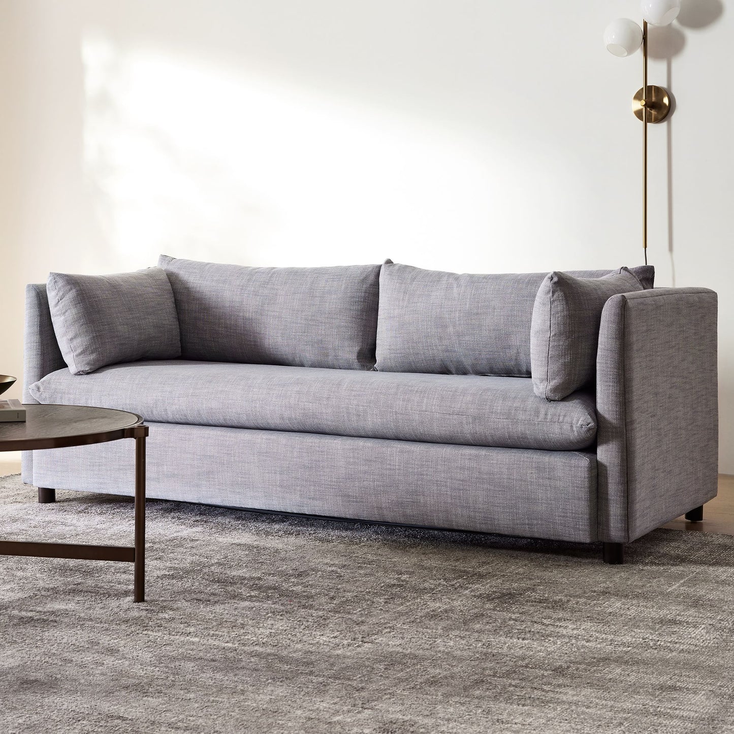 A modern living room showcases the Shelter Sleeper Sofa with three back cushions, accompanied by a round wooden coffee table on a textured gray rug. In the corner against a white wall stands a brass floor lamp with globe-shaped lights.