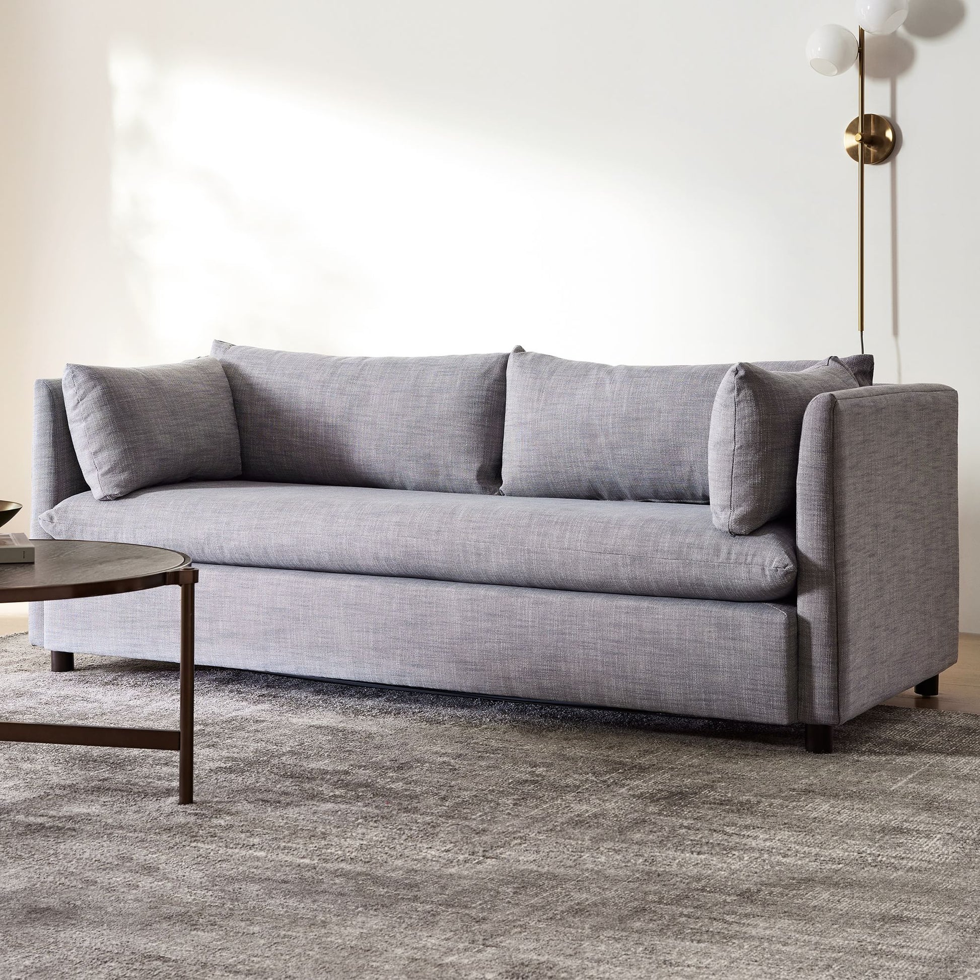 A modern living room showcases the Shelter Sleeper Sofa with three back cushions, accompanied by a round wooden coffee table on a textured gray rug. In the corner against a white wall stands a brass floor lamp with globe-shaped lights.