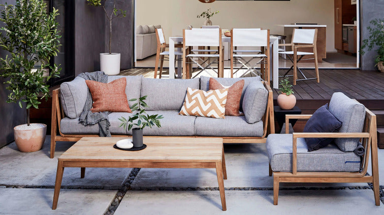 A stylish outdoor patio setting featuring a wooden sofa and armchair with gray cushions and patterned throw pillows. A matching wooden coffee table is in front of the seating. The background shows a dining area with a table and chairs, and some potted plants.