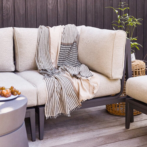 An outdoor seating area with cream-colored cushions and a black-and-white checkered blanket draped over the side. A small potted plant and wicker basket sit on the wooden floor. A table with a bowl of grapes is partially visible in the foreground.