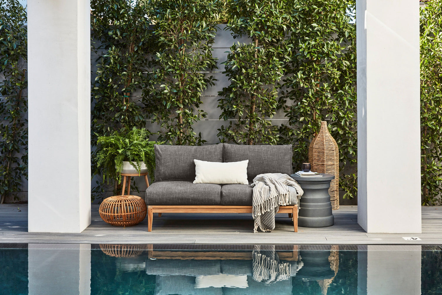 A modern outdoor patio setup with a gray cushioned wooden sofa, white and gray pillows, a gray throw, a side table, wicker accessories, and greenery in the background. In front of the sofa is a swimming pool with reflections of the furniture in the water.