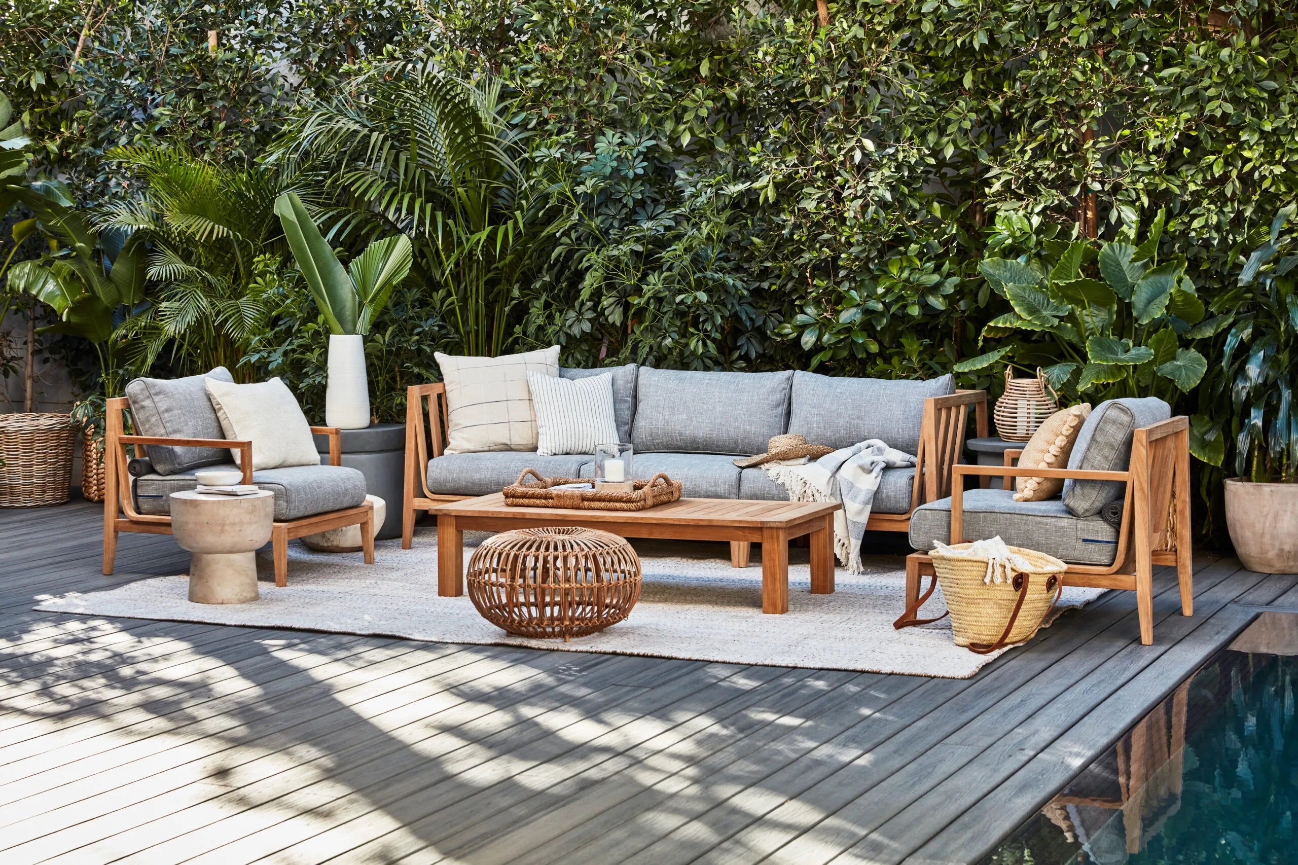 An inviting outdoor patio features a wooden furniture set with grey cushions, including a sofa and two chairs. A wooden coffee table, a woven basket, and various potted plants surround the seating area. The patio overlooks a lush garden with a wooden deck.