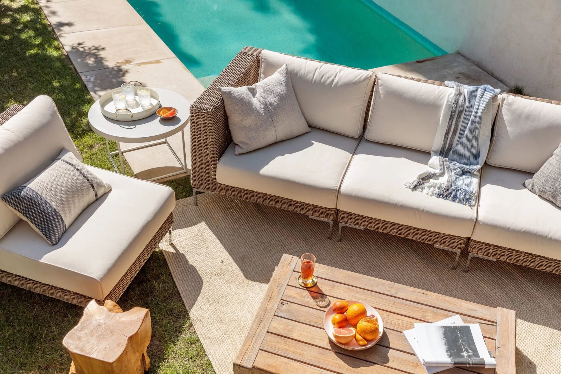 A sunlit outdoor seating area next to a swimming pool, featuring a wicker sofa and armchair with cream cushions and gray pillows. A wooden coffee table holds a bowl of peaches, a glass of juice, and a magazine. A cozy throw blanket drapes over one arm of the sofa.