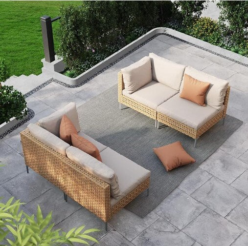 A patio with gray tiles features two beige cushioned wicker sofas on a gray rug. Two orange pillows are on one sofa, and one orange pillow is on the ground. Green grass, bushes, and trees surround the area.