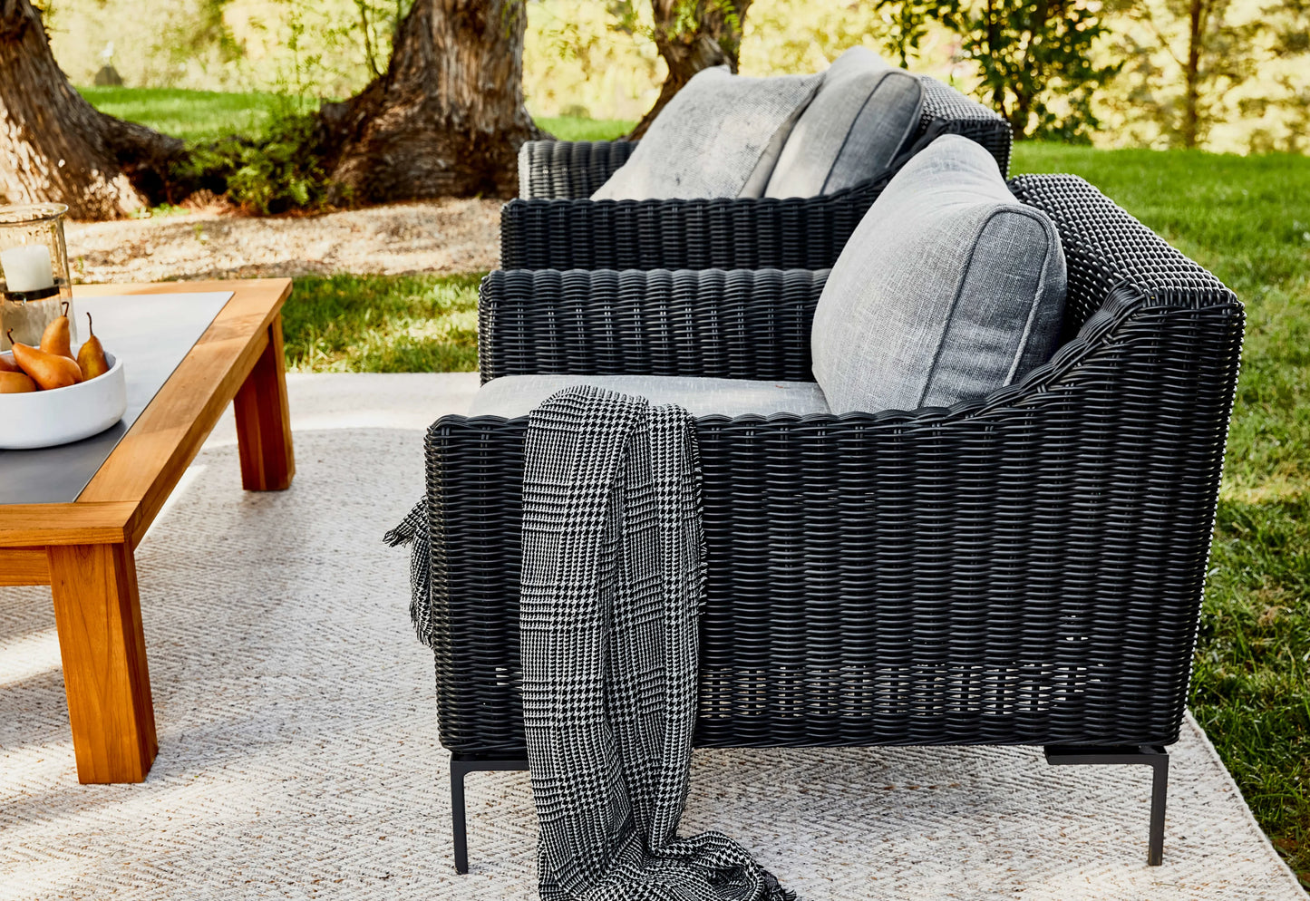 A cozy outdoor seating area includes a black wicker outdoor sofa and armchairs set in a five-seat configuration, complete with gray cushions. A plaid blanket is draped over one of the armchairs. Positioned on a beige rug is a wooden coffee table, adorned with a candle and tray of fruit, all nestled among green trees and grass.