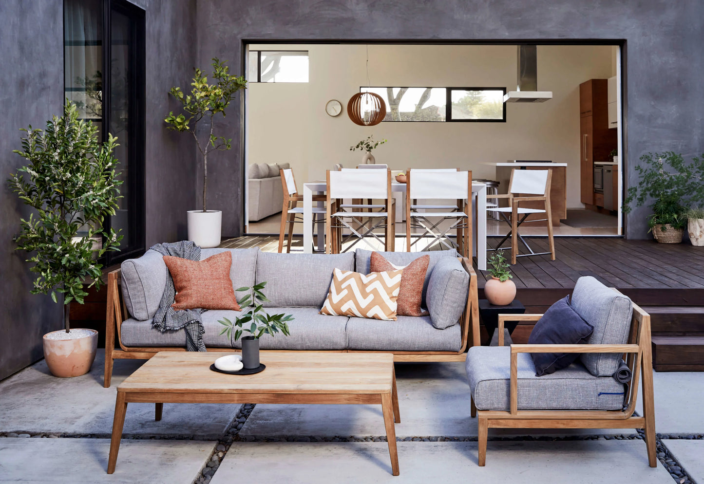 A contemporary patio showcases a Teak Outdoor Sofa with Armless Chairs - Five Seat complete with gray cushions and orange pillows, complemented by a wooden coffee table and potted plants. In the background, a dining area equipped with high chairs and a large window provides a glimpse into the interior.