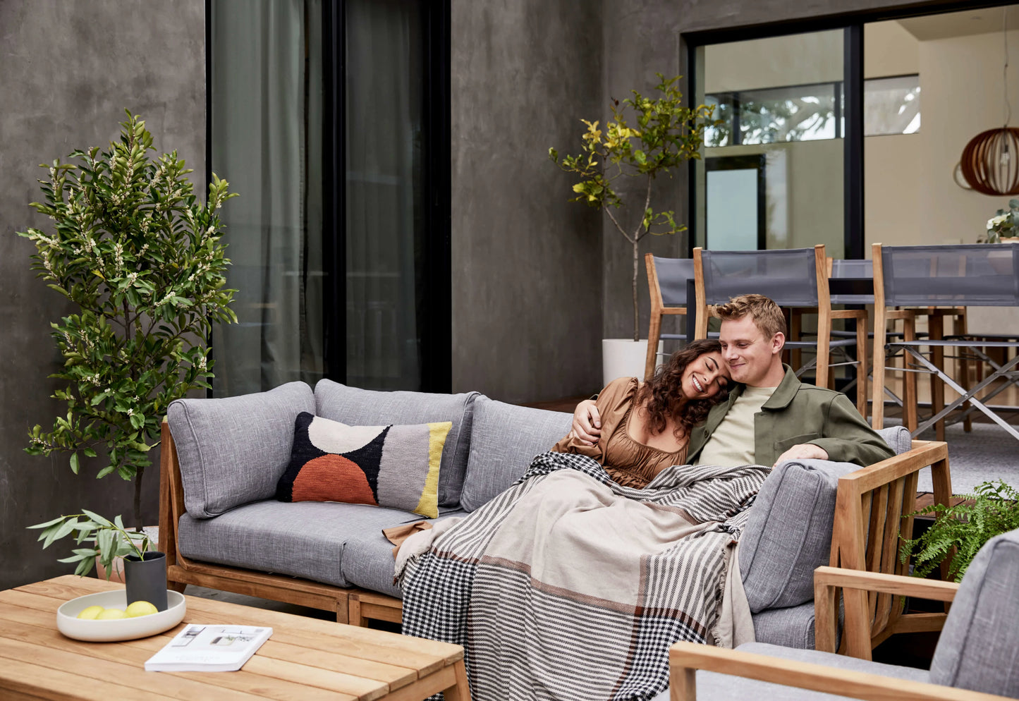 A couple sits cozily on the Teak Outdoor Sofa with Armless Chairs - Five Seat, wrapped in a blanket. They're surrounded by plants and contemporary furniture, with a table holding a bowl and magazine in the foreground, and a dining set in the background.