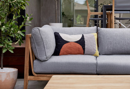 A modern outdoor patio showcases a Teak Outdoor Sofa with Armless Chairs - Five Seat, featuring wooden accents and adorned with a patterned cushion in black, white, orange, and yellow hues. A wooden table graces the foreground while a potted plant is positioned on the left.