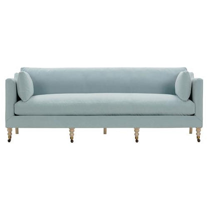 A French grey sofa with a modern and sleek design takes center stage in the image, featuring straight arms and two cylindrical side pillows. This Madeline sofa stands on brown wooden legs equipped with small brass casters for added mobility.