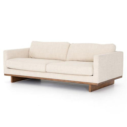 The Ethan Modern Classic Cream Performance Cushion Back Sofa features a sleek design in beige, complemented by two cushions and a low wooden base, all set against a white background.