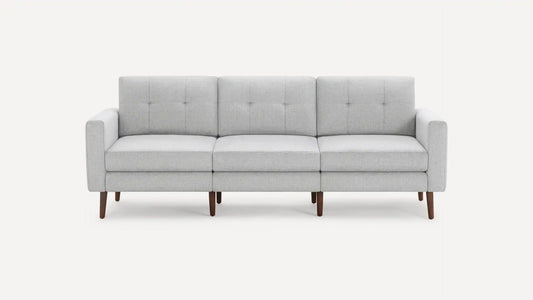 The Nomad Fabric Sofa is a contemporary gray piece with a minimalist design, featuring three cushions and elegant wooden legs, set against a simple backdrop.