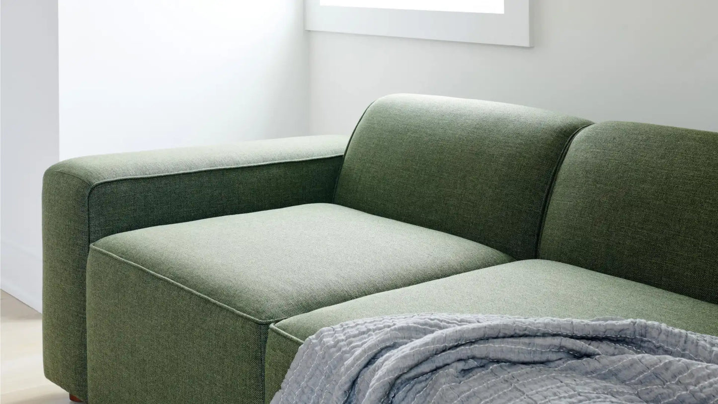 The Mambo Loveseat, featuring a modern green design with minimalist flair, is placed in the corner of a bright room. A soft, textured gray blanket is elegantly draped across its cushion, fostering a cozy and inviting ambiance.