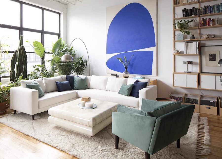 A modern living room with a white sectional sofa and green armchair, surrounded by various green plants. A large, abstract blue painting dominates the wall. Bookshelves and a floor lamp are also present. Natural light pours in from large windows, illuminating the space.