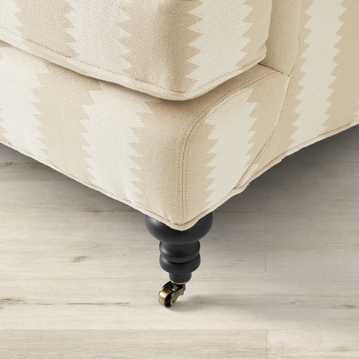 Close-up of the corner of the Sanderson x Giles Deacon Bedford 87" Sofa in beige upholstery featuring a chevron pattern. The sofa is supported by dark wooden legs with brass caster wheels, placed on a light wood floor.