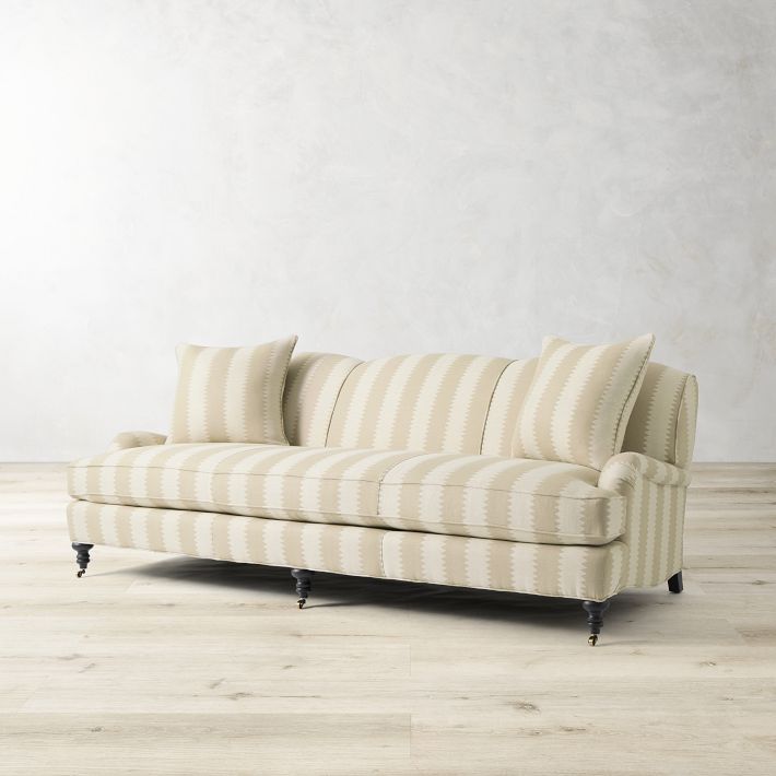 The Sanderson x Giles Deacon Bedford 87" Sofa, featuring a beige striped design and curved armrests, complete with two matching cushions, is placed on a light wooden floor in front of a plain, light-colored wall.