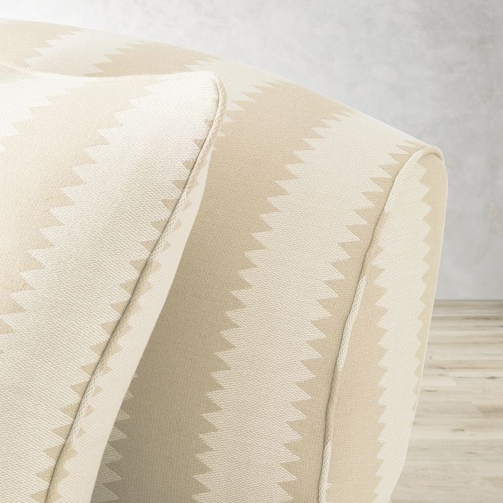 Close-up of the arm of the Sanderson x Giles Deacon Bedford 87" Sofa, showcasing its beige chevron zigzag pattern with alternating light and dark shades. In the background, a light gray wall complements the wooden floor.