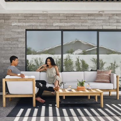 A man and a woman relax on the Haven Sectional, a modern outdoor patio set with white cushions and wooden frames, positioned against a brick wall. In front of them is a coffee table adorned with a potted plant and glass jars. Large windows reflect the image of a white building and trees.