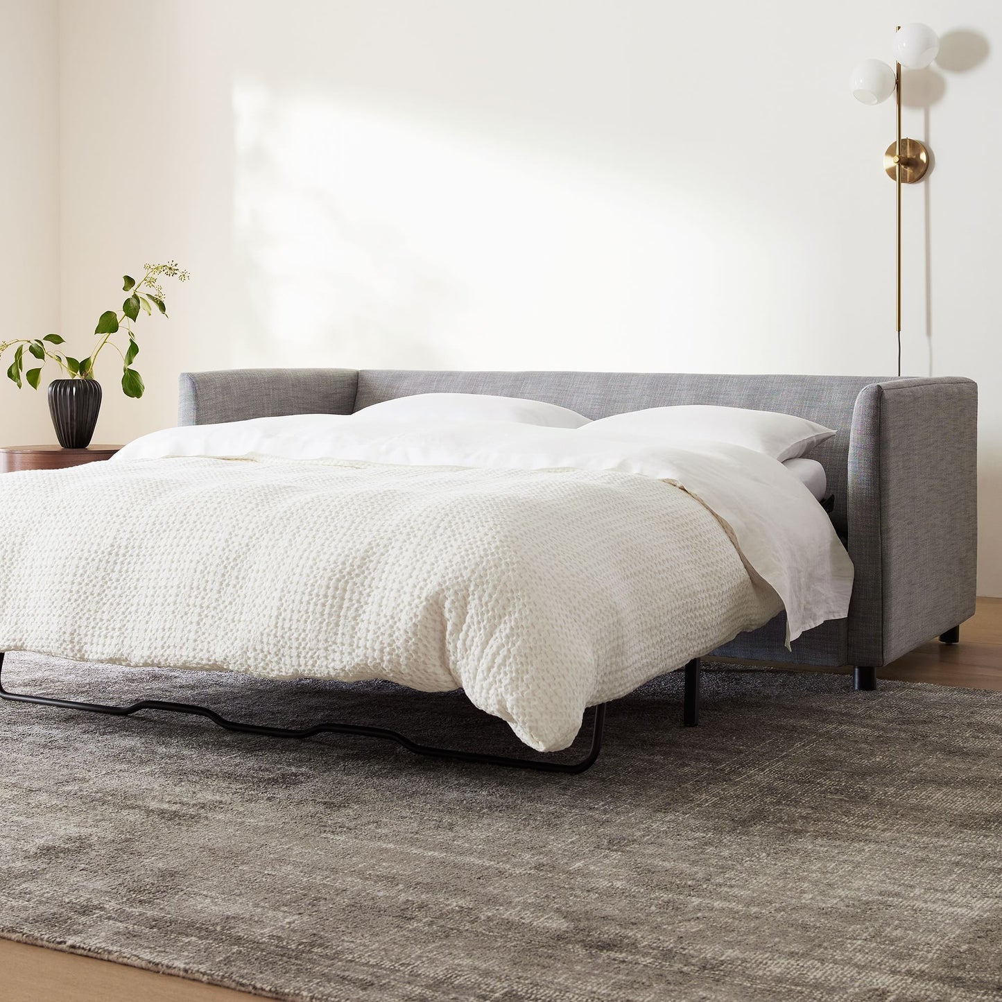 A modern living room features the Shelter Sleeper Sofa in its extended form, adorned with white bedding. Nearby, a green potted plant is perched on a wooden side table. The floor boasts a soft, textured gray rug, while a wall-mounted lamp contributes an elegant touch.