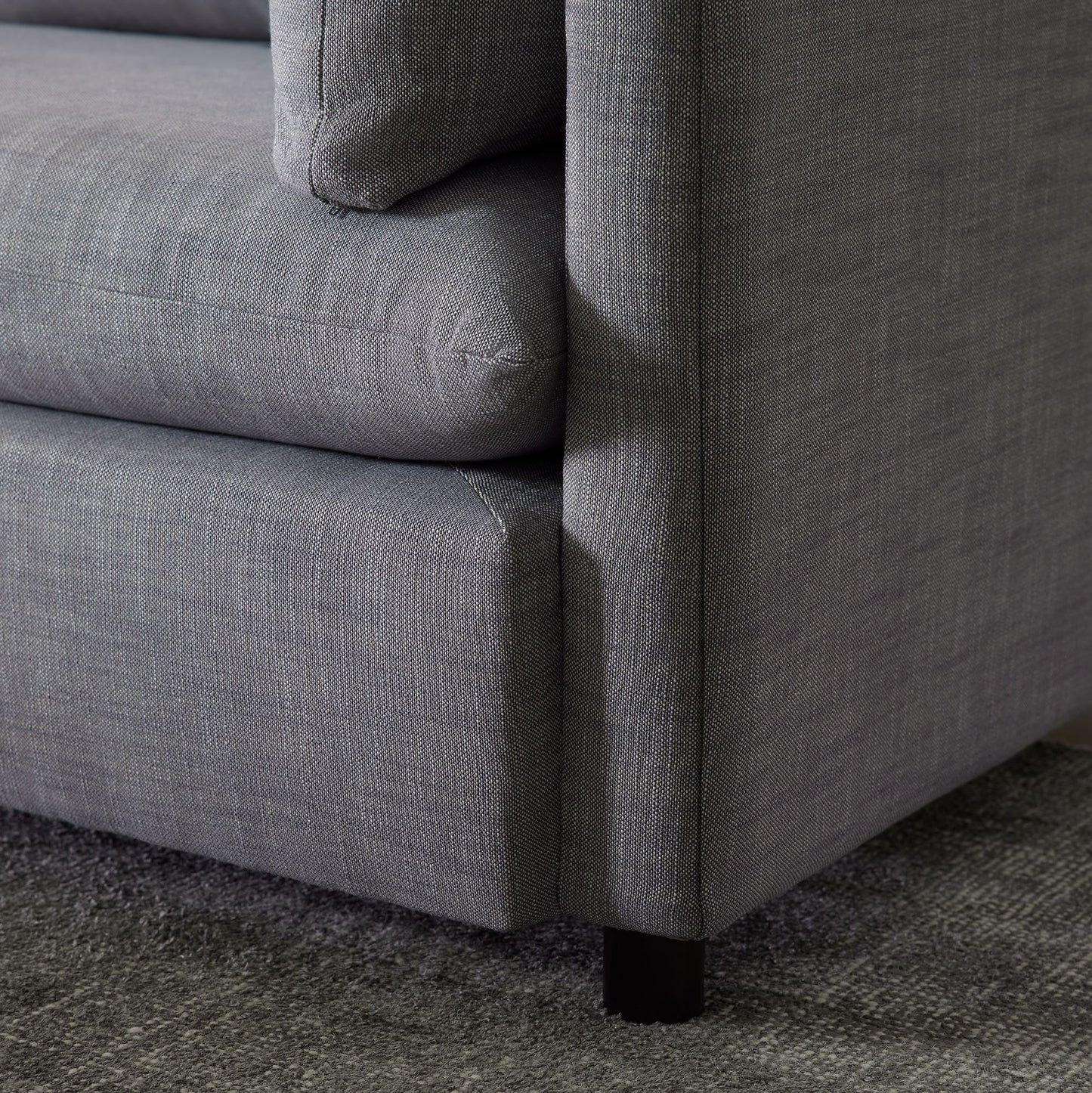 A close-up view of the Shelter Sleeper Sofa's corner reveals the fabric's texture and a sleek black leg. Positioned on a matching gray carpet, it exemplifies modern design with its clean lines.