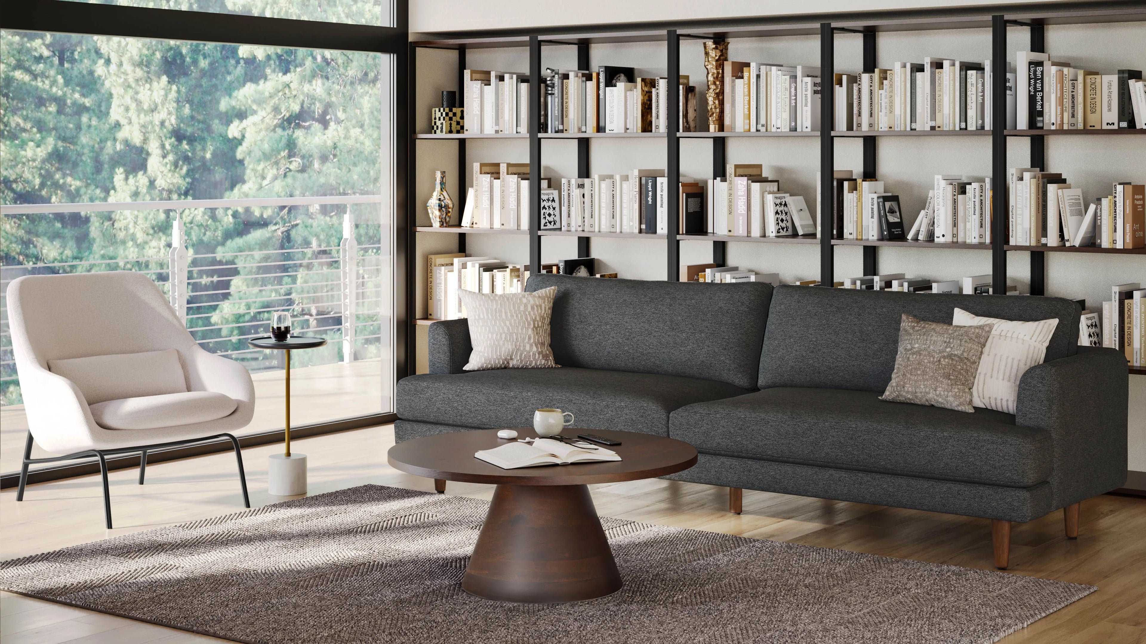 A modern living room features a dark grey sofa with multiple cushions, a round wooden coffee table with books and a cup on it, a white armchair by the window, and a large bookshelf filled with books. Glass doors open to a balcony with a view of trees.