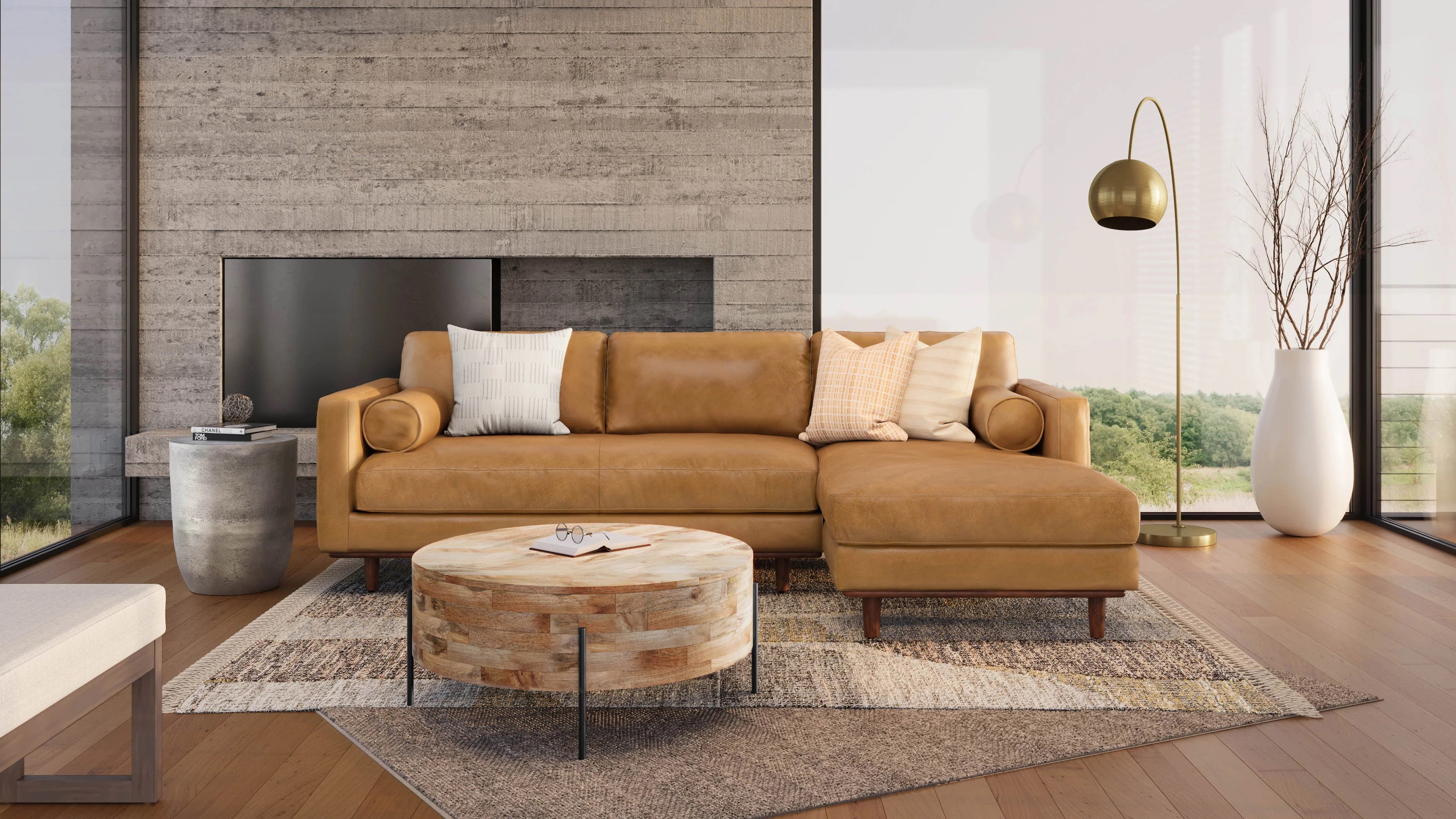 A modern living room featuring a brown leather sectional sofa with pillows, a round wooden coffee table, and a floor lamp with a gold finish. The room has wooden floors, a large window with a scenic view, and a gray-textured wall with a built-in fireplace.