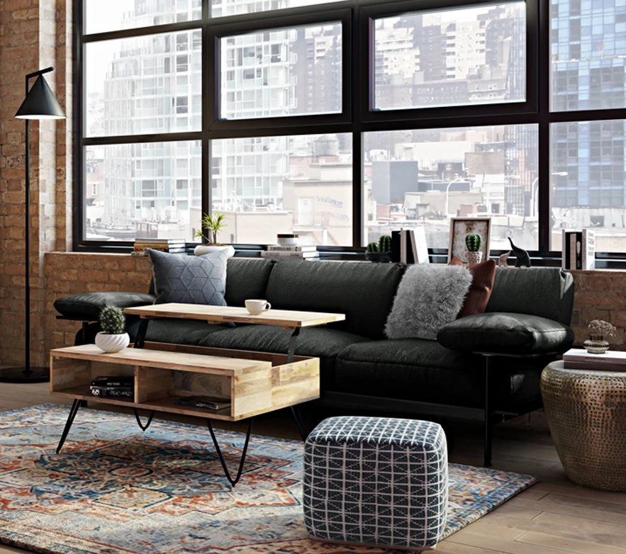 A modern living room with a large window overlooking a cityscape. It features a black sofa with grey and white pillows, a wooden coffee table with an open shelf, a patterned rug, a geometric pouf, a metal floor lamp, and exposed brick walls.