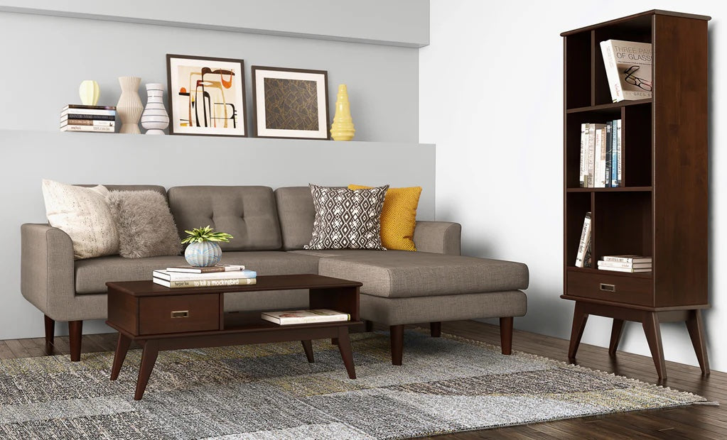 A modern living room features a gray sectional sofa adorned with various pillows, a wooden coffee table with books and a small plant, a tall wooden bookshelf with books and decor items, and a light gray rug. The walls are decorated with framed artwork and vases.