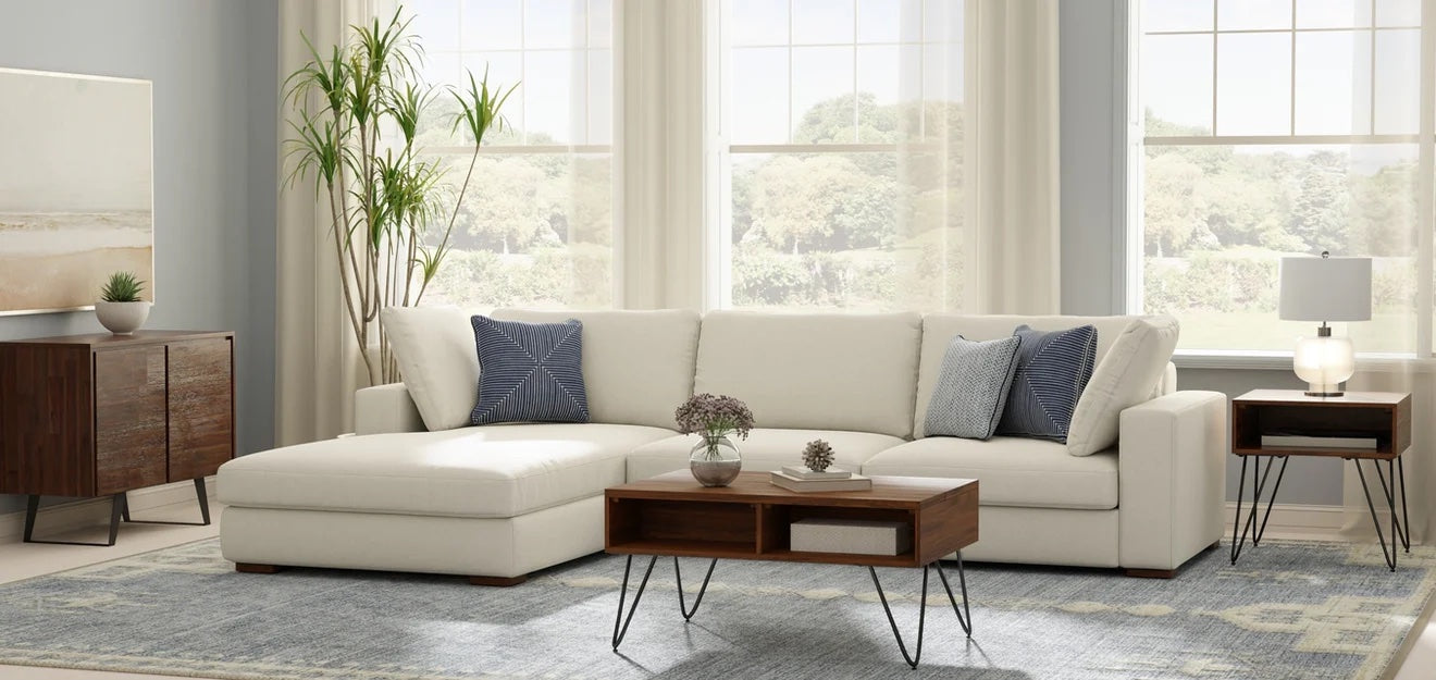 A modern living room with a white sectional sofa, adorned with blue and gray pillows. A wooden coffee table with metal legs is centered on a blue patterned rug. A tall plant and a wooden cabinet flank the scene, with large windows in the background letting in natural light.