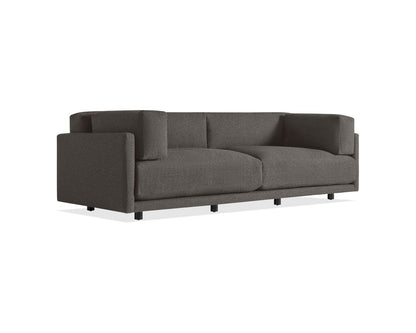 The Sunday 102-Inch Sofa is a modern piece with a boxy design, crafted from dark gray fabric and featuring plush seat and back cushions. Its straight armrests and short, black legs contribute to its sleek and contemporary appearance.