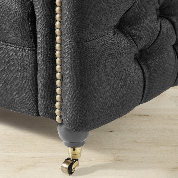 Close-up of the Soho Sofa in dark gray upholstery, showcasing button-style detailing and decorative bronze studs along the edge. The sofa is set on a light wooden floor and is equipped with a wheel caster.