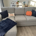 A cozy living room scene featuring the Playday Beige 93'' 2 Piece Left Arm Facing Pop-Up Sleeper Chaise Sectional with USB & USB-C Port. The sectional is decorated with a black blanket, blue and pumpkin-shaped pillows, and a decorative pillow that reads "Hello Pumpkin." A dining table can be seen in the background.