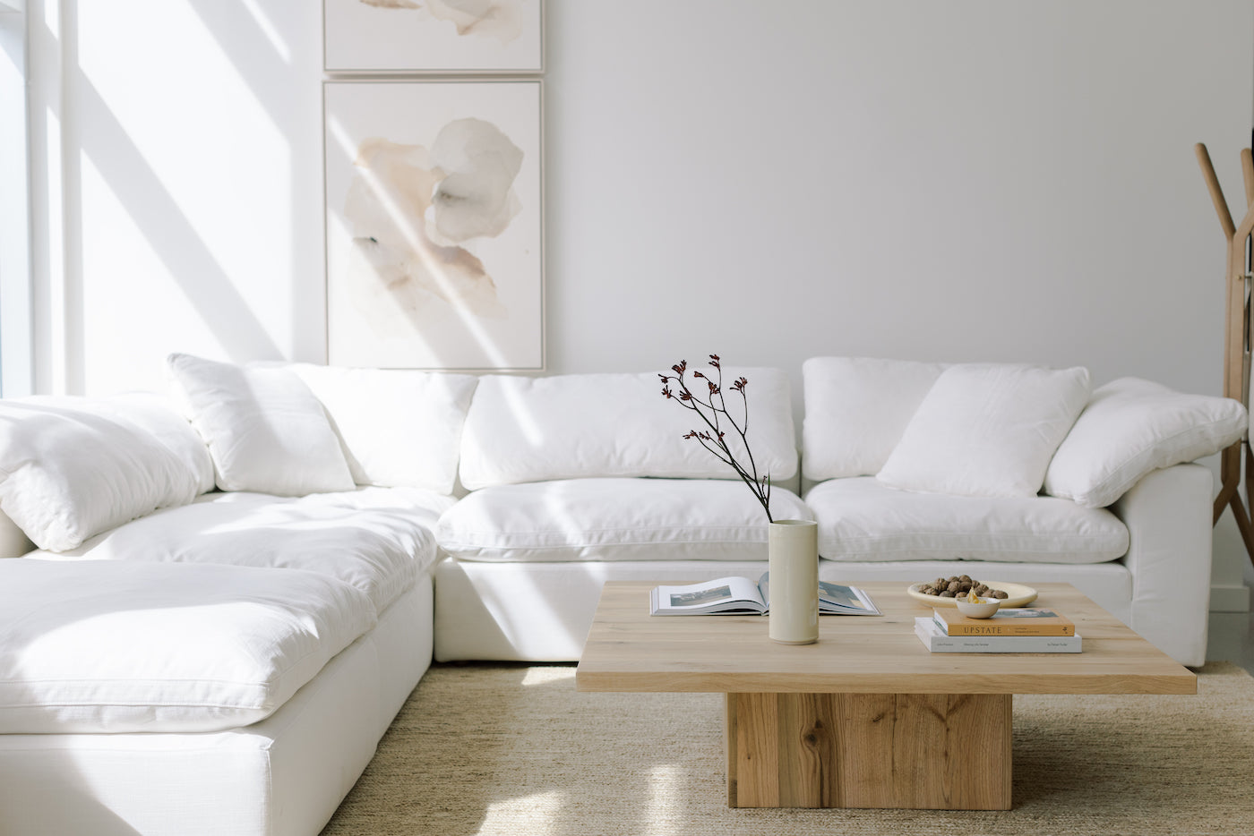 Minimalist living room with a large white sectional sofa, wooden coffee table, and neutral decor. A small vase with branches sits on the table alongside books. Two abstract art pieces are on the wall, and soft natural light filters through the space.