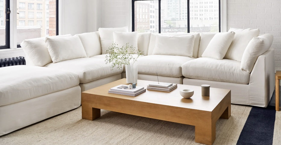 A spacious living room features a white fabric sectional sofa with plush cushions. A light wood coffee table in front holds decorative items, including a vase with greenery, stacked books, and small bowls. Large windows offer a view of buildings outside.