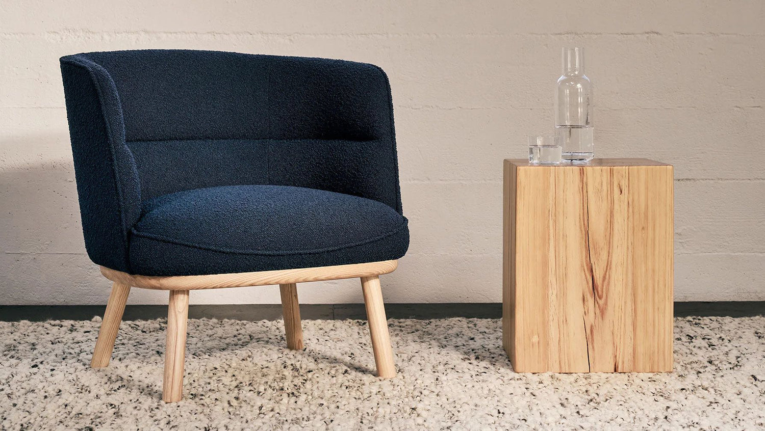 A modern black upholstered chair with wooden legs is positioned next to a minimalist wooden side table. On the table, there is a clear glass bottle and a glass of water. The setup is placed on a light textured carpet against a beige wall.