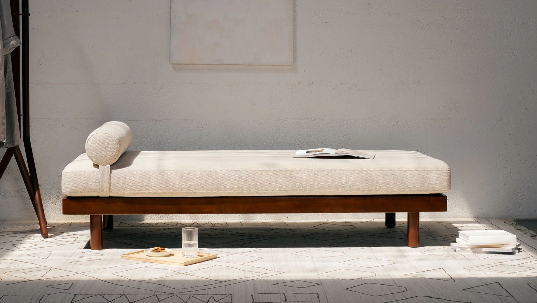 A minimalist daybed with beige upholstery and a cylindrical pillow is placed in a bright room with a white wall. A small wooden tray with a glass of water and biscuits rests on the floor nearby, along with an open book on the daybed. Geometric rug lies beneath.